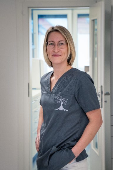 Frau mit blonden Haaren in beruflicher Kleidung, lächelt in einem hellen Raum.